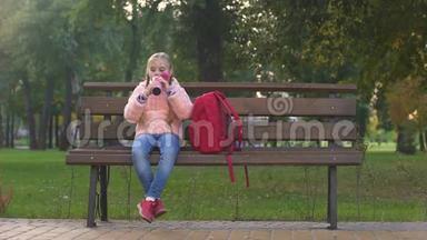 快乐的女学生拿着背包，一边坐在长凳上，一边从<strong>热水</strong>杯里喝茶